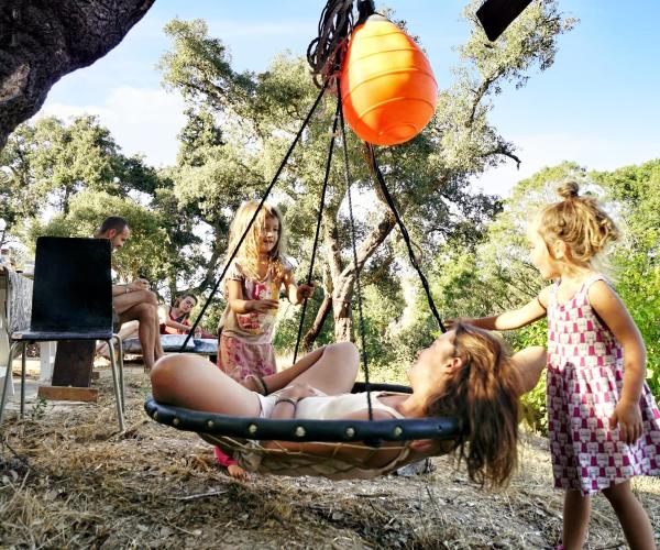 A parent on a hanging swing with children playing around.